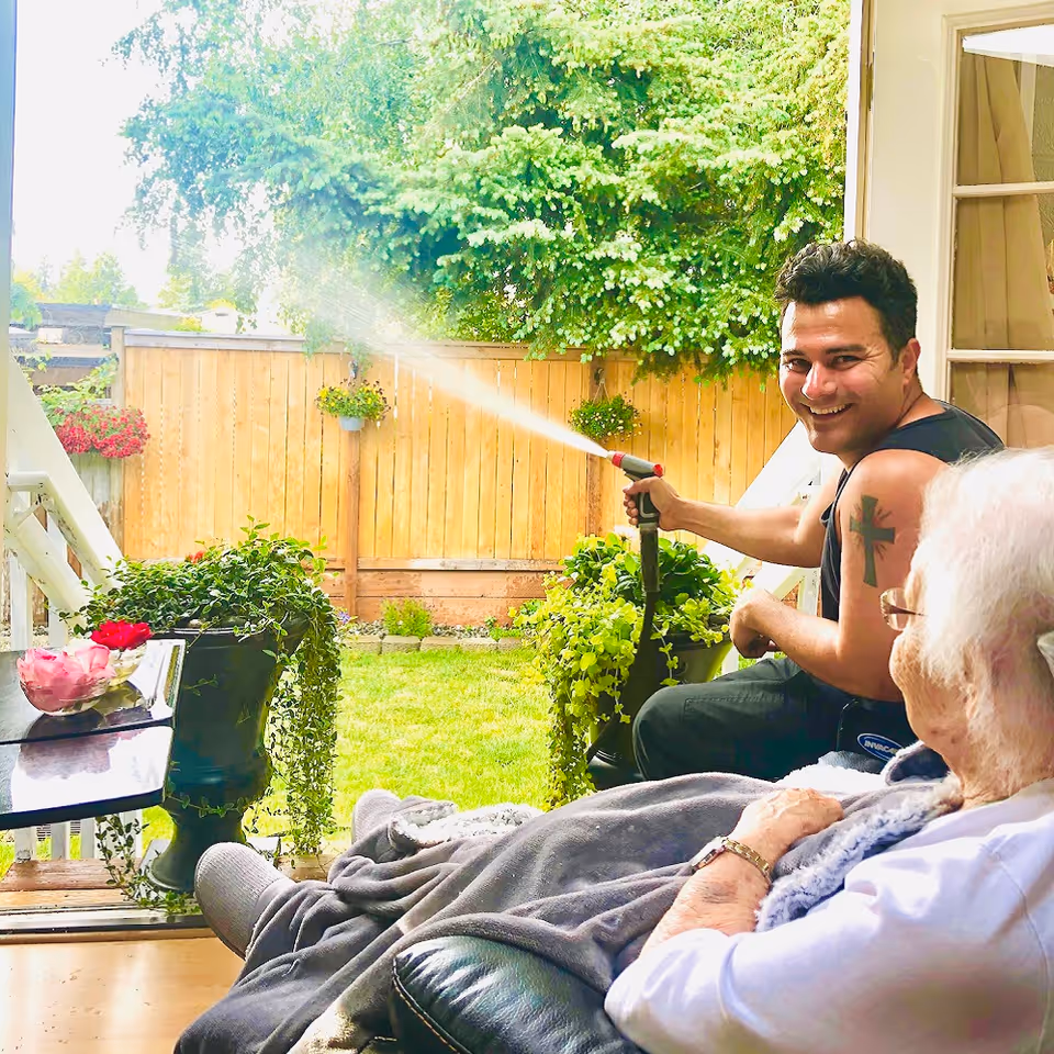 A young man smiling while watering plants with a garden hose on a porch. An elderly person with white hair and glasses is sitting nearby, covered with a blanket, watching him. The scene is set in a backyard with a wooden fence, green grass, and hanging flower pots.