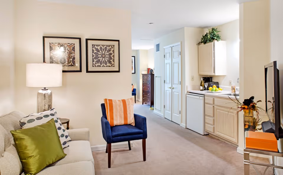 Bright living area with a sofa, blue armchair, lamp and wall art, opening to a small kitchenette down a short hallway.