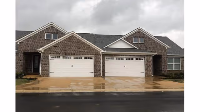 Front exterior view of a residential building with two attached garages, brick walls, and a wet driveway under an overcast sky.