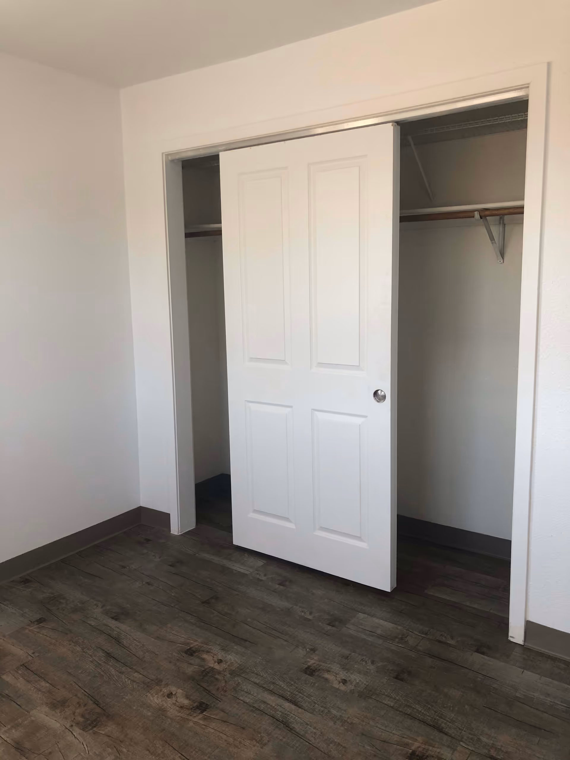 An empty room with wood-patterned flooring and white walls featuring a partially open white sliding closet door revealing an empty closet with a hanging rod and shelf.