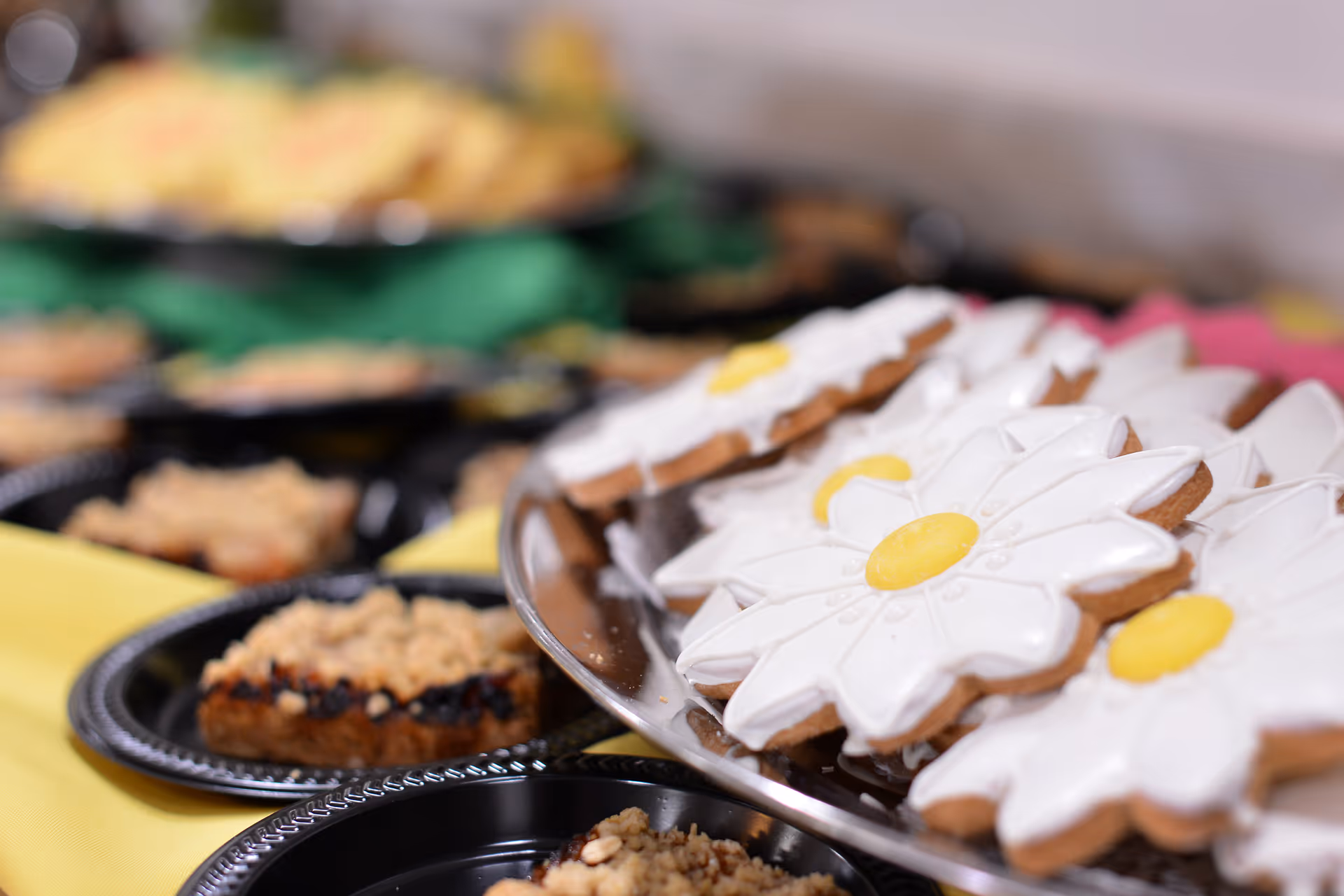 Close-up view of a tray with flower-shaped cookies decorated with white icing and yellow centers, surrounded by plates with crumb-topped dessert bars on a table with a yellow tablecloth.