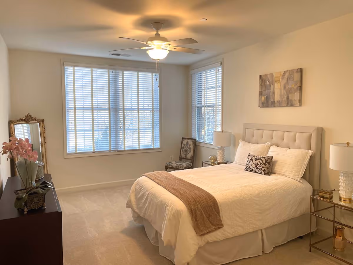 A bright bedroom with a large window covered by white blinds. The room features a neatly made bed with white bedding, a beige tufted headboard, and a brown throw blanket. There are two bedside tables with matching white lamps, a decorative chair near the window, a dresser with a pink orchid plant and a large ornate mirror leaning against the wall. A ceiling fan with a light is mounted on the ceiling.