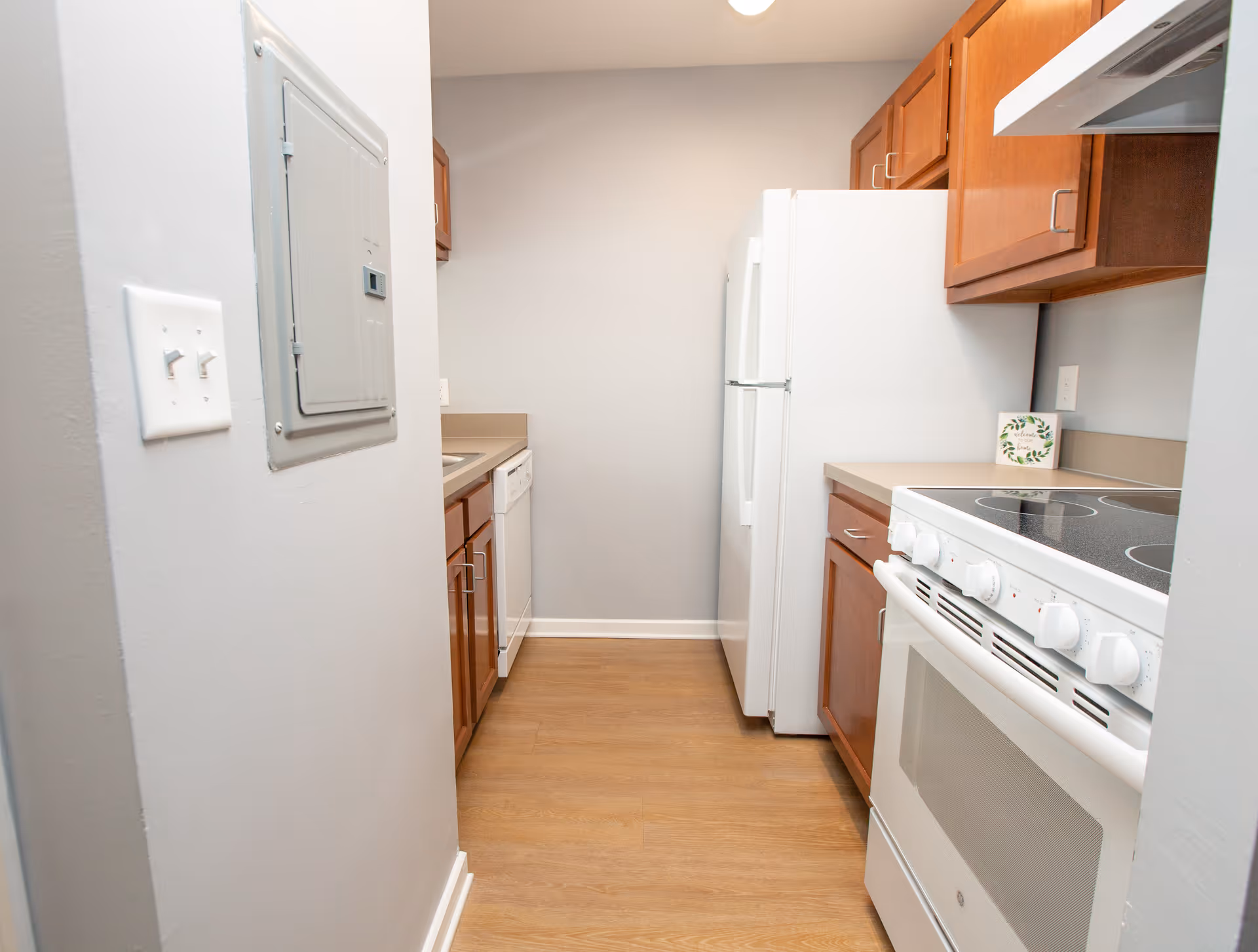 A narrow kitchen with wooden cabinets, a white electric stove, a white refrigerator, a dishwasher, and a sink. The floor is wooden, and the walls are painted light gray.