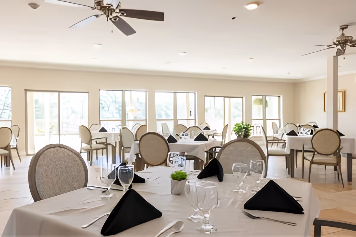 Bright dining room with several tables set with white tablecloths, black napkins, glassware and chairs beneath ceiling fans and large windows.