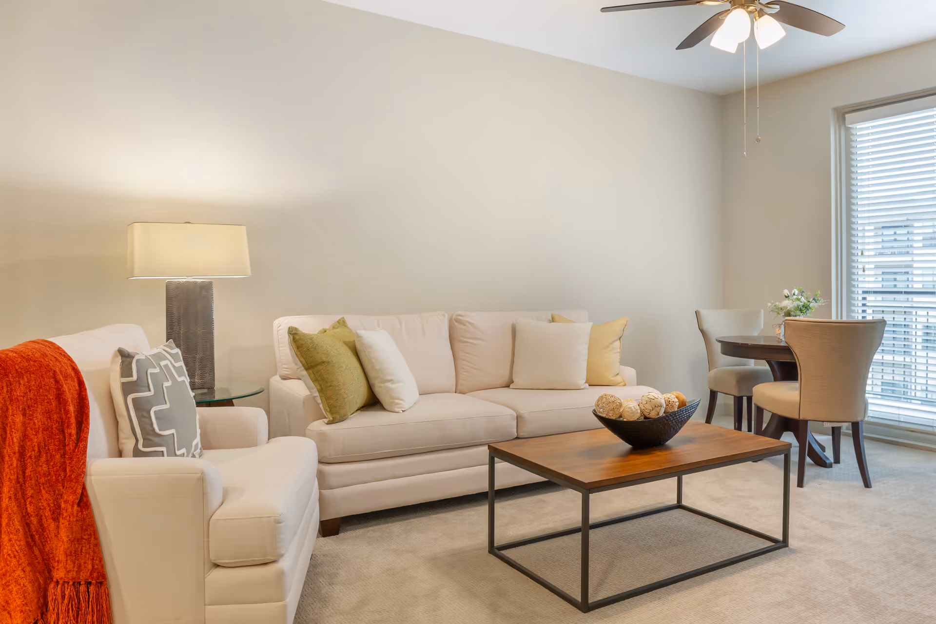 A bright living room with a beige sofa and armchair, decorated with green and white pillows. A wooden coffee table with a decorative bowl sits in front of the sofa. A floor lamp with a rectangular shade is next to the armchair, which has an orange throw draped over it. In the background, there is a small round dining table with four beige upholstered chairs near a large window with blinds. A ceiling fan with lights is visible above.