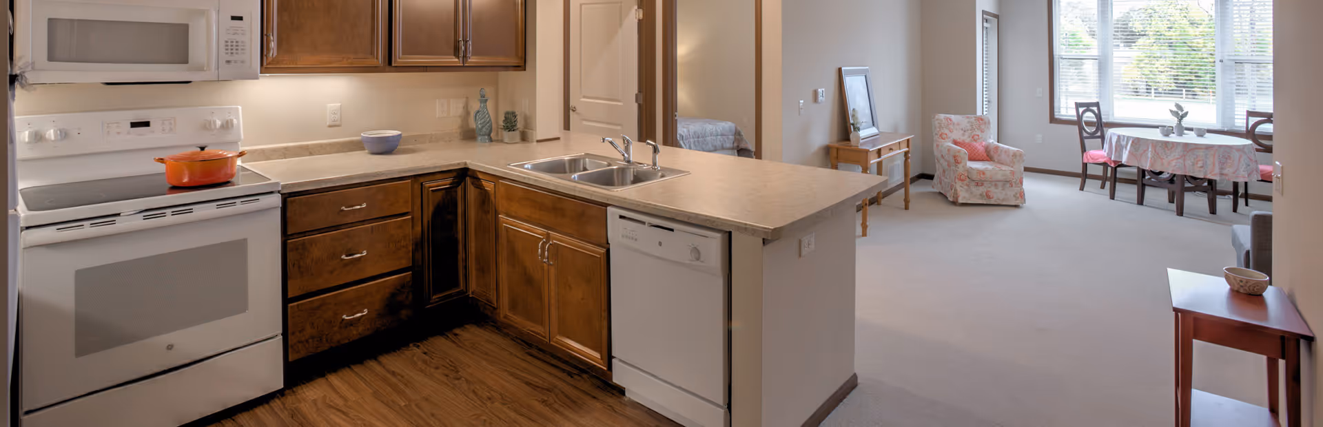 Open kitchen with white appliances, wooden cabinets and an island overlooking a carpeted living and dining area with large windows.