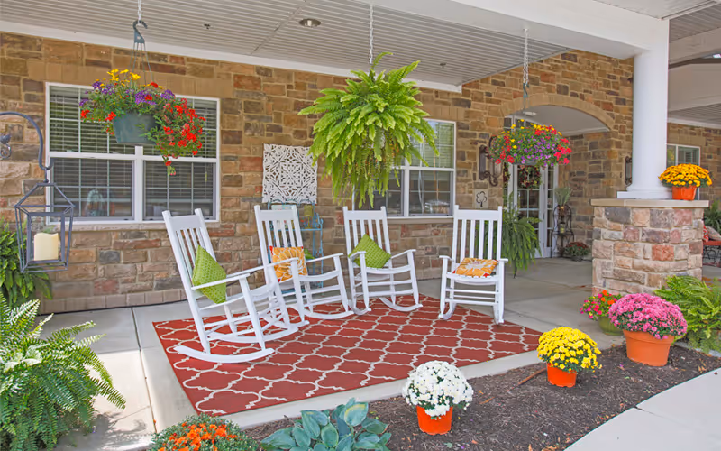 Covered outdoor patio area with four white rocking chairs arranged on a red patterned rug. The patio is decorated with hanging flower baskets, potted plants, and green ferns. The background features a stone wall with windows and a white column supporting the roof.