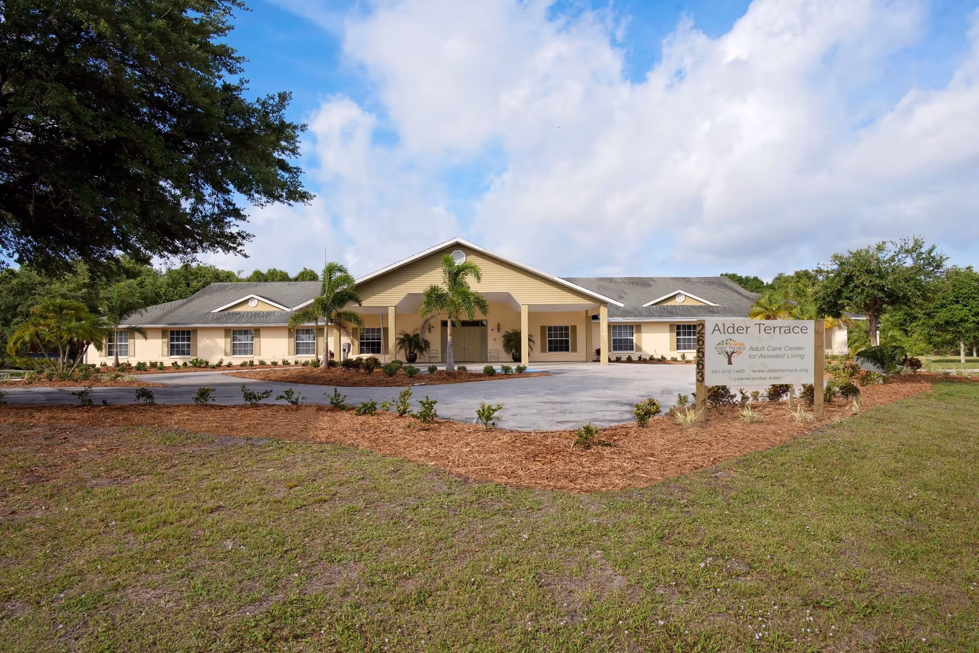 Front exterior view of Alder Terrace Adult Care Center, a single-story building with a beige facade, multiple windows, and a covered entrance. The building is surrounded by landscaped greenery, palm trees, and a clear blue sky with some clouds.
