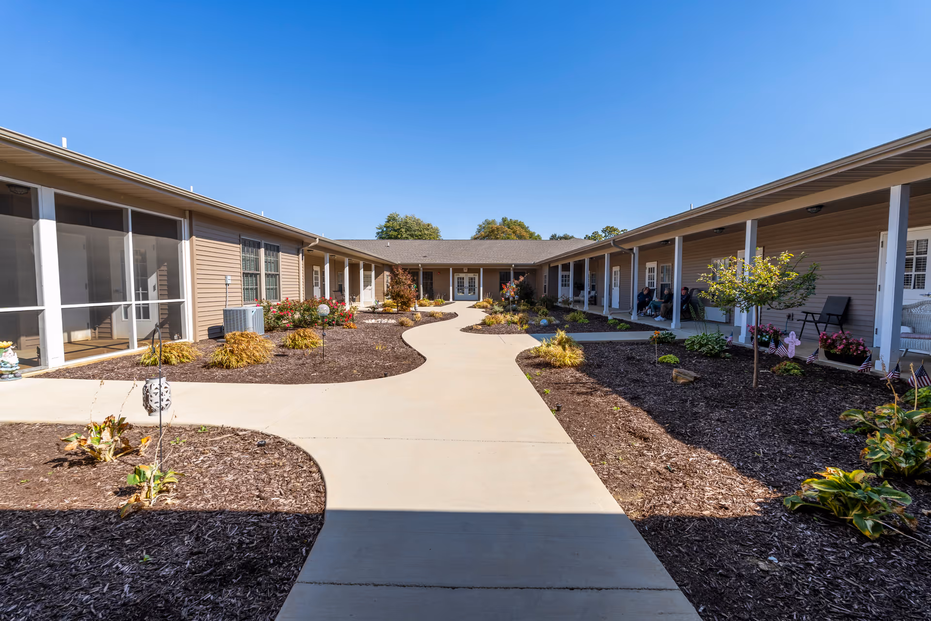 Sunlit courtyard with a paved walkway, landscaped beds, and single-story residential wings with covered porches.