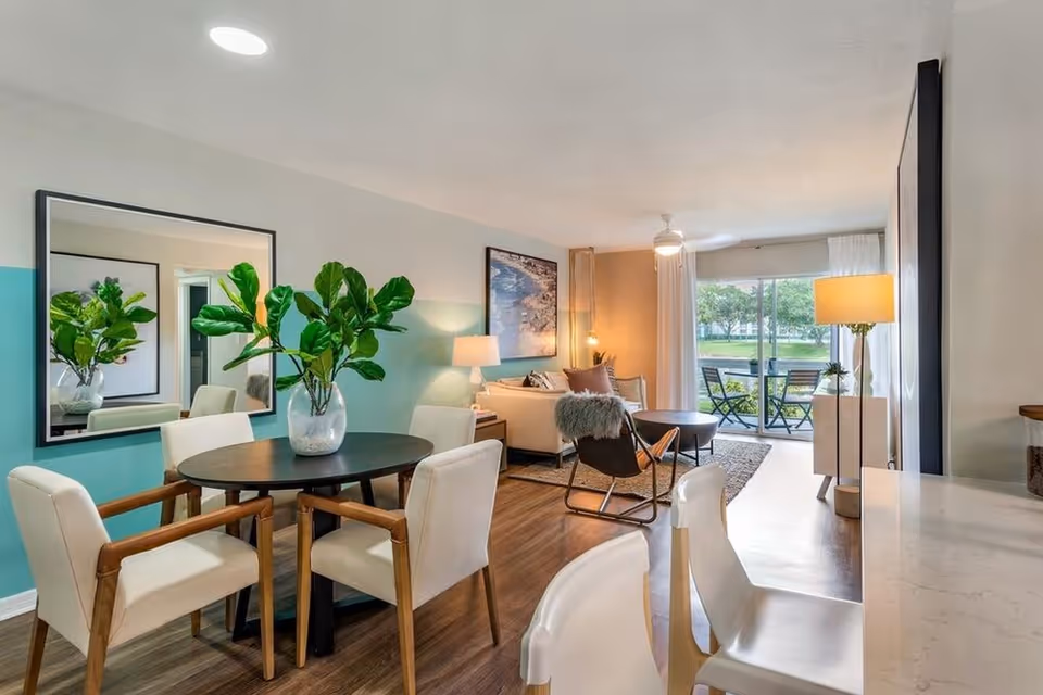 A modern living and dining area with a round black dining table surrounded by four white cushioned chairs with wooden arms. A large mirror hangs on the teal accent wall behind the table, reflecting a green plant in a glass vase on the table. The living room features a beige sofa, a black chair with a fur throw, a round coffee table, and a floor lamp. Large sliding glass doors open to an outdoor patio with chairs and a table, overlooking a green lawn and trees.