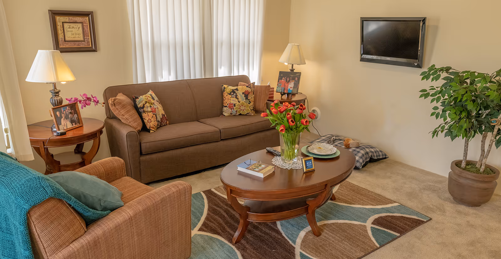 A cozy living room with a brown sofa adorned with floral and solid pillows, a matching armchair with a green throw blanket and pillow, a wooden oval coffee table with a vase of red tulips, books, and a small framed picture. Two side tables each hold a lamp and framed family photos. A flat-screen TV is mounted on the wall above a potted plant, and a colorful area rug covers part of the beige carpeted floor.