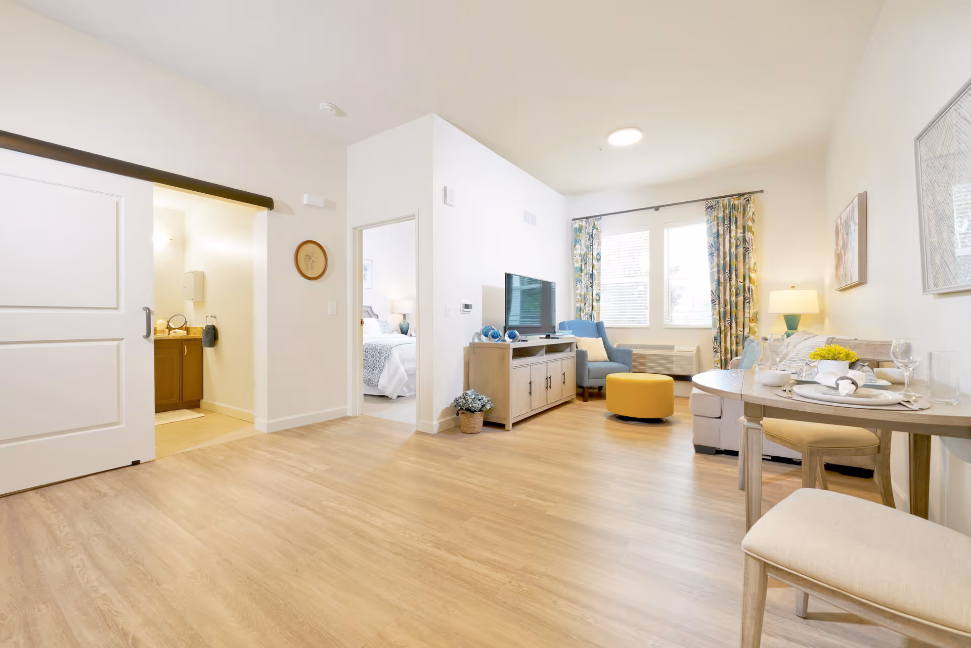 Bright and spacious senior living apartment interior featuring a living room with a TV on a wooden stand, a blue armchair, a yellow ottoman, and a beige sofa. A round dining table with two chairs is set with plates, glasses, and a small flower arrangement. Large windows with floral curtains allow natural light to fill the room. A doorway leads to a bedroom with a neatly made bed, and another doorway reveals a bathroom with a vanity and mirror. The flooring is light wood, and the walls are painted white.