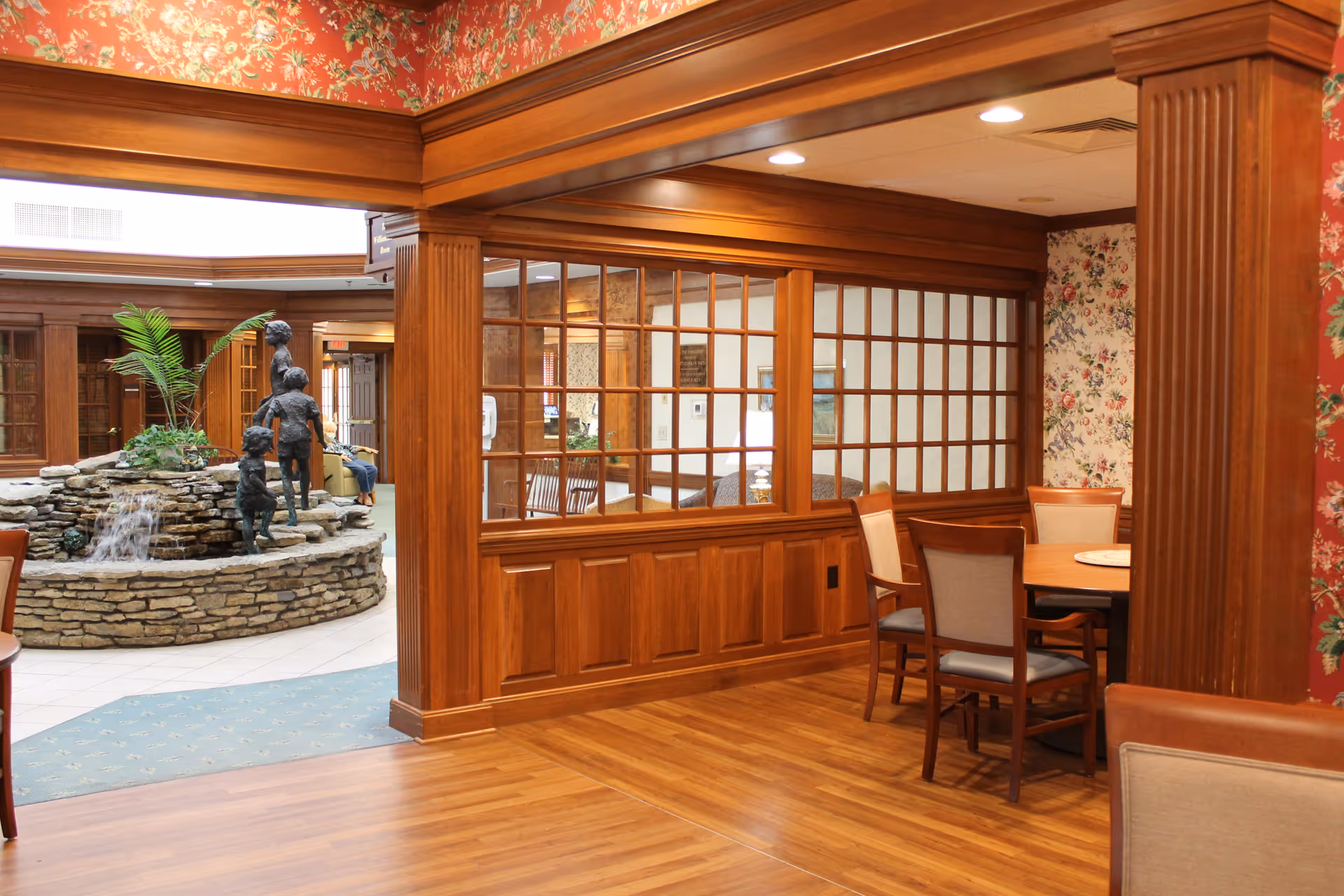 Interior view of a retirement community featuring a wooden floor and wood-paneled walls with large windows. There is a round table with chairs in a corner, floral wallpaper, and a stone water fountain with a bronze sculpture of children playing near the center. The area is well-lit with ceiling lights.