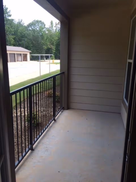A small covered balcony or patio area with a black metal railing overlooking a landscaped area with mulch and small plants. In the background, there is a parking lot and a building with garage doors, surrounded by trees.