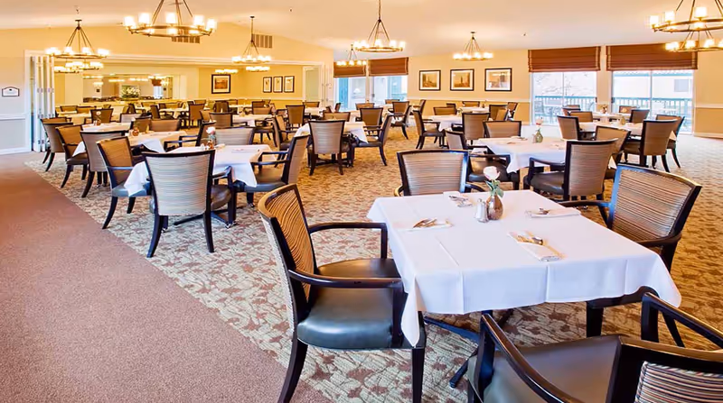 A spacious dining room with multiple tables covered in white tablecloths, each set with silverware and a small vase with a flower. The room has patterned carpet, wooden chairs with cushioned seats, and warm lighting from chandeliers hanging from the ceiling. Large windows allow natural light to enter the room.