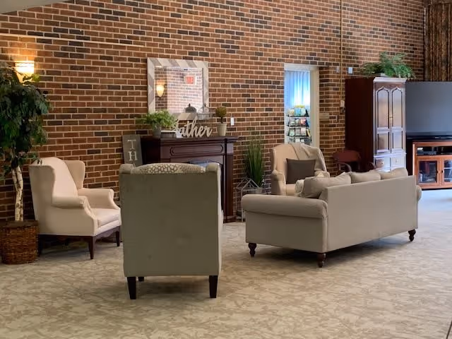 A cozy living room area with beige and light gray upholstered chairs and sofas arranged around a fireplace with a mirror and decorative plants on the mantel. The room has a brick wall, carpeted floor, a large wooden cabinet with a TV, and a potted plant in the corner.