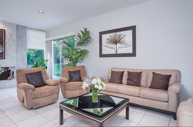 Bright living room with a beige sofa, two recliner chairs, a glass-top coffee table with flowers, and wall art.