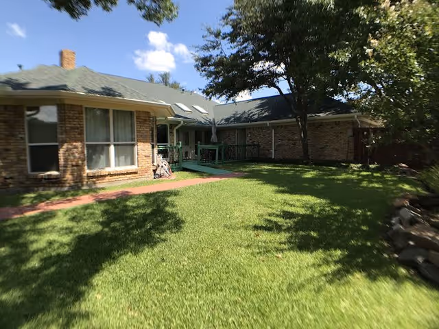 Single-story brick assisted living building with a green lawn, walkway, shaded porch, and a walker near the entrance.