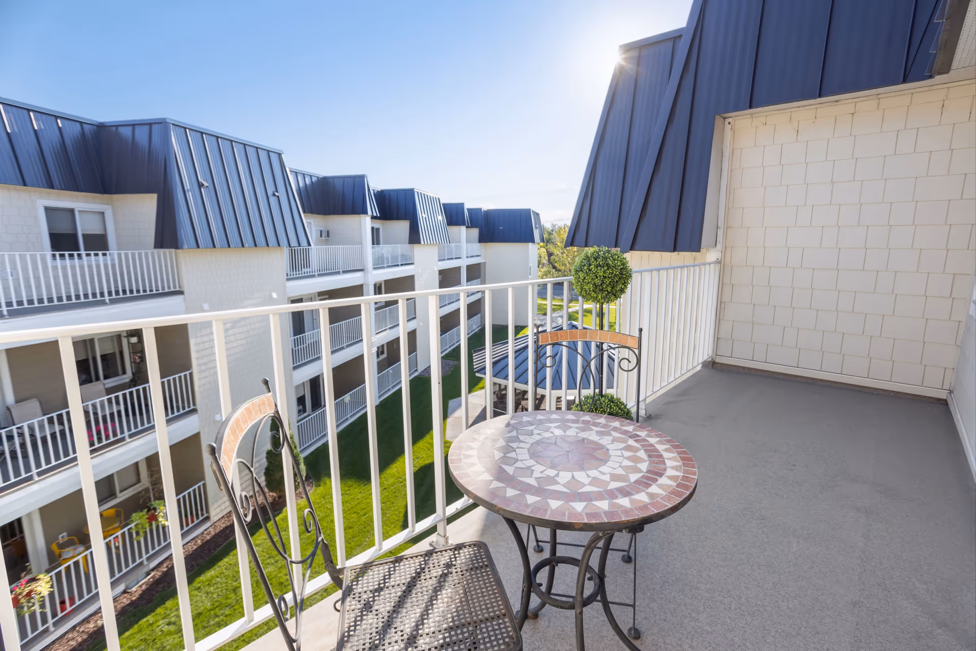 A balcony with a small round mosaic table and two metal chairs with wooden accents. The balcony overlooks a courtyard with green grass and a multi-story building with white railings and dark blue roofs under a clear blue sky.