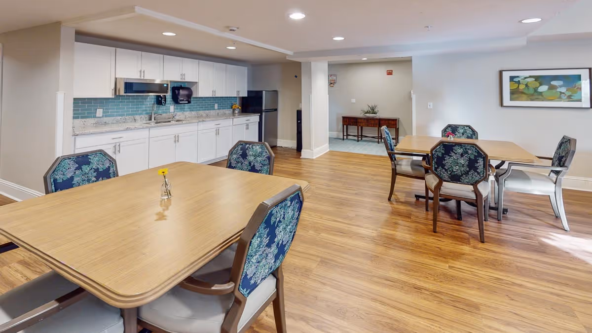 Bright common dining area with wood floors, multiple tables and upholstered chairs, and a white kitchenette with a blue tile backsplash.