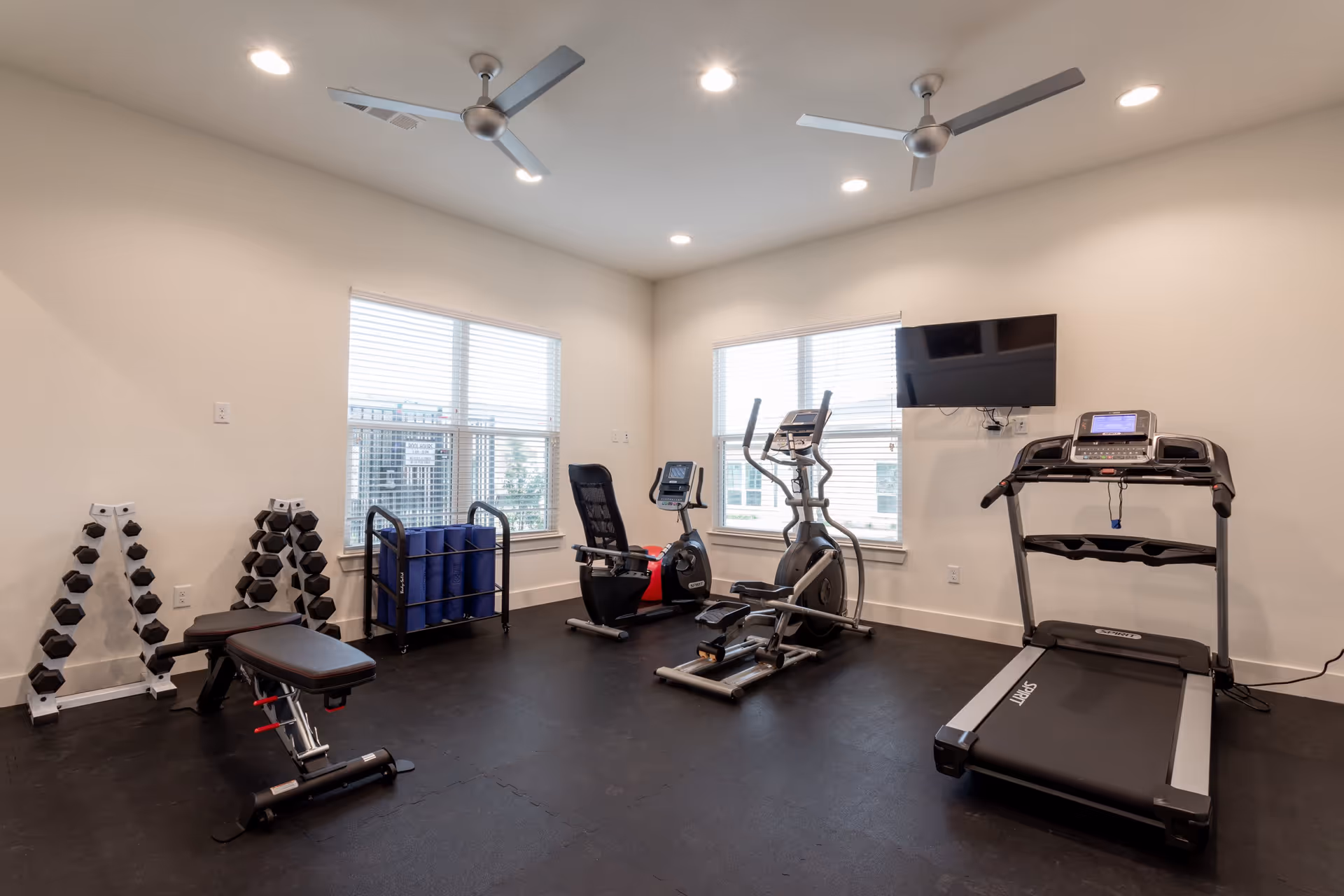 Small fitness room with a treadmill, elliptical, recumbent bike, weight bench, dumbbells and wall-mounted TV.