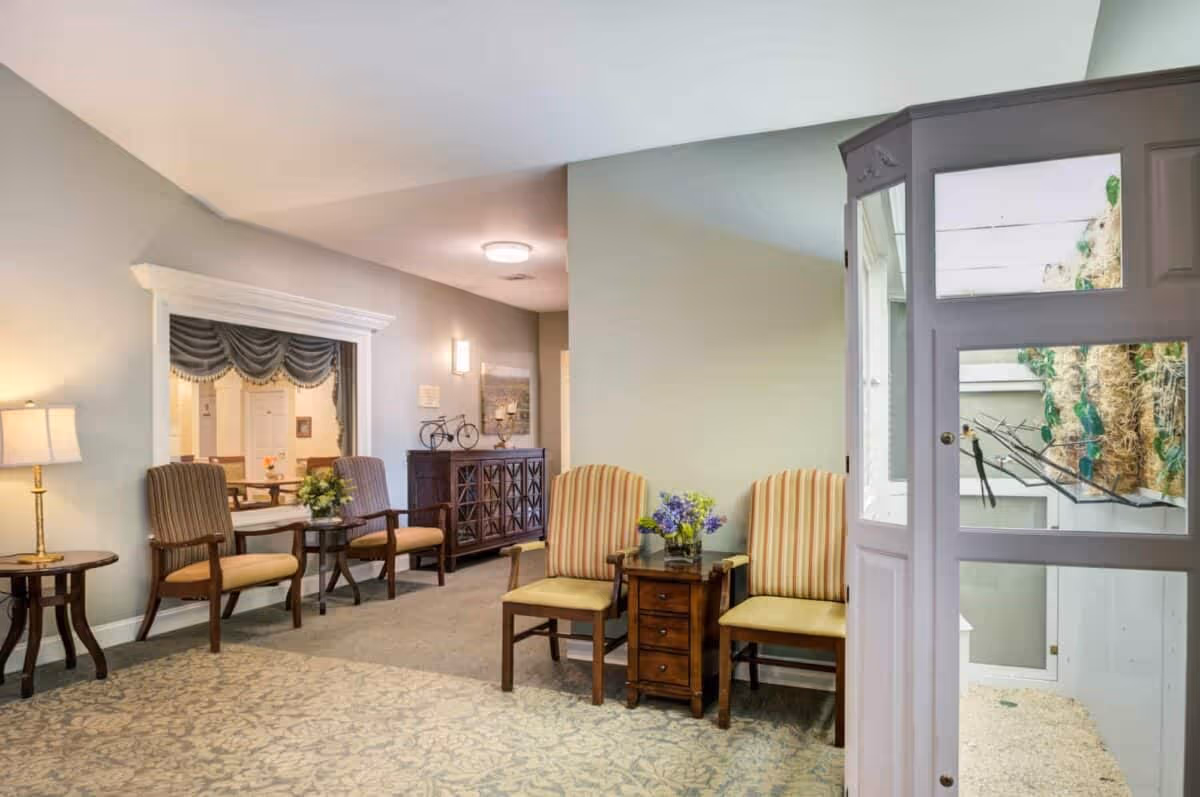 A cozy interior common area with four wooden chairs featuring striped cushions arranged around two small wooden tables with flower arrangements. A lamp on a side table is on the left, and a window with a decorative curtain looks into another room with tables and chairs. The walls are painted in soft neutral tones, and there is a cabinet with decorative items along the back wall. On the right side, there is a white enclosure with glass panels containing a birdcage with birds inside.