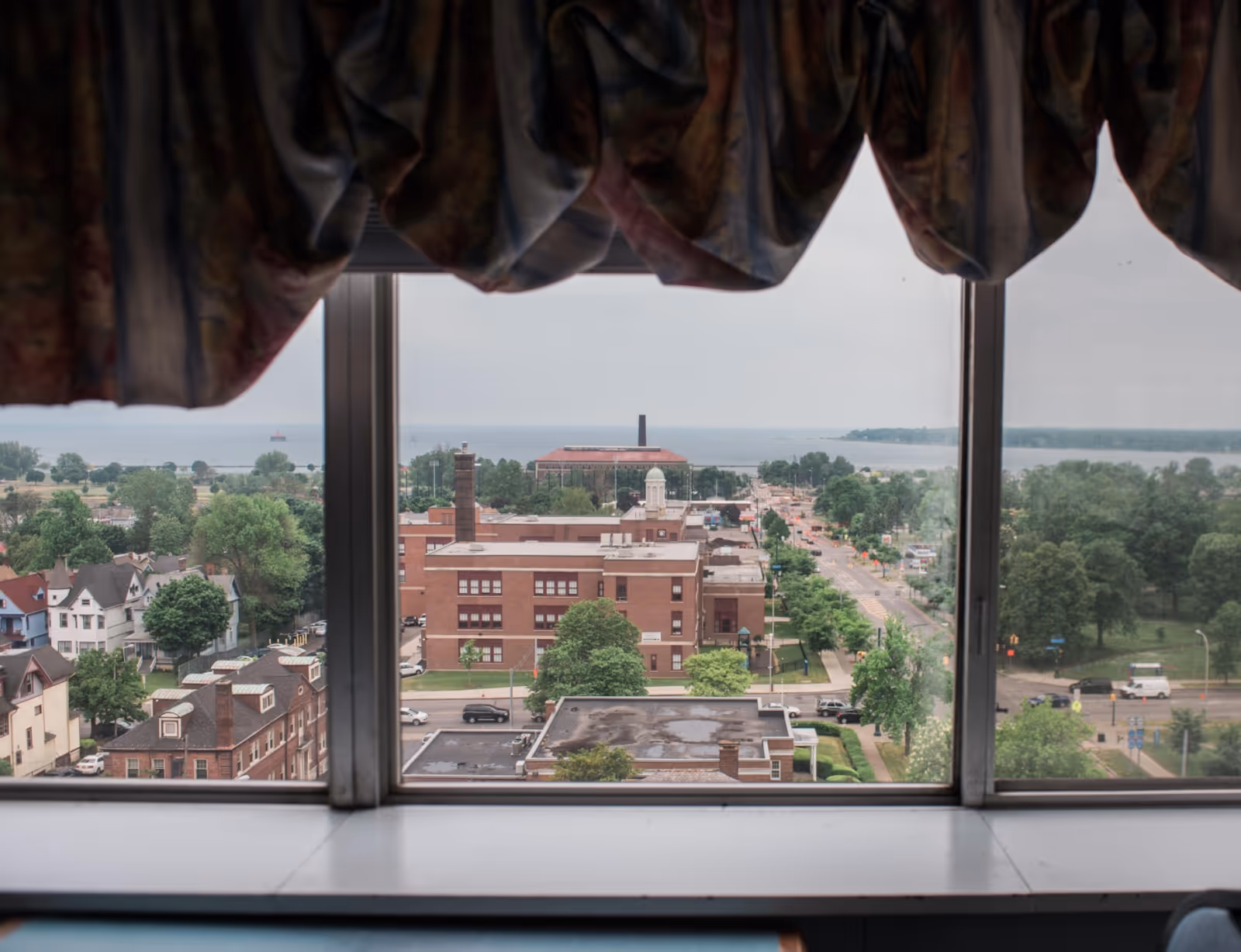 View through a window with floral curtains looking out over a town with buildings, trees, roads, and a body of water in the distance under a cloudy sky.