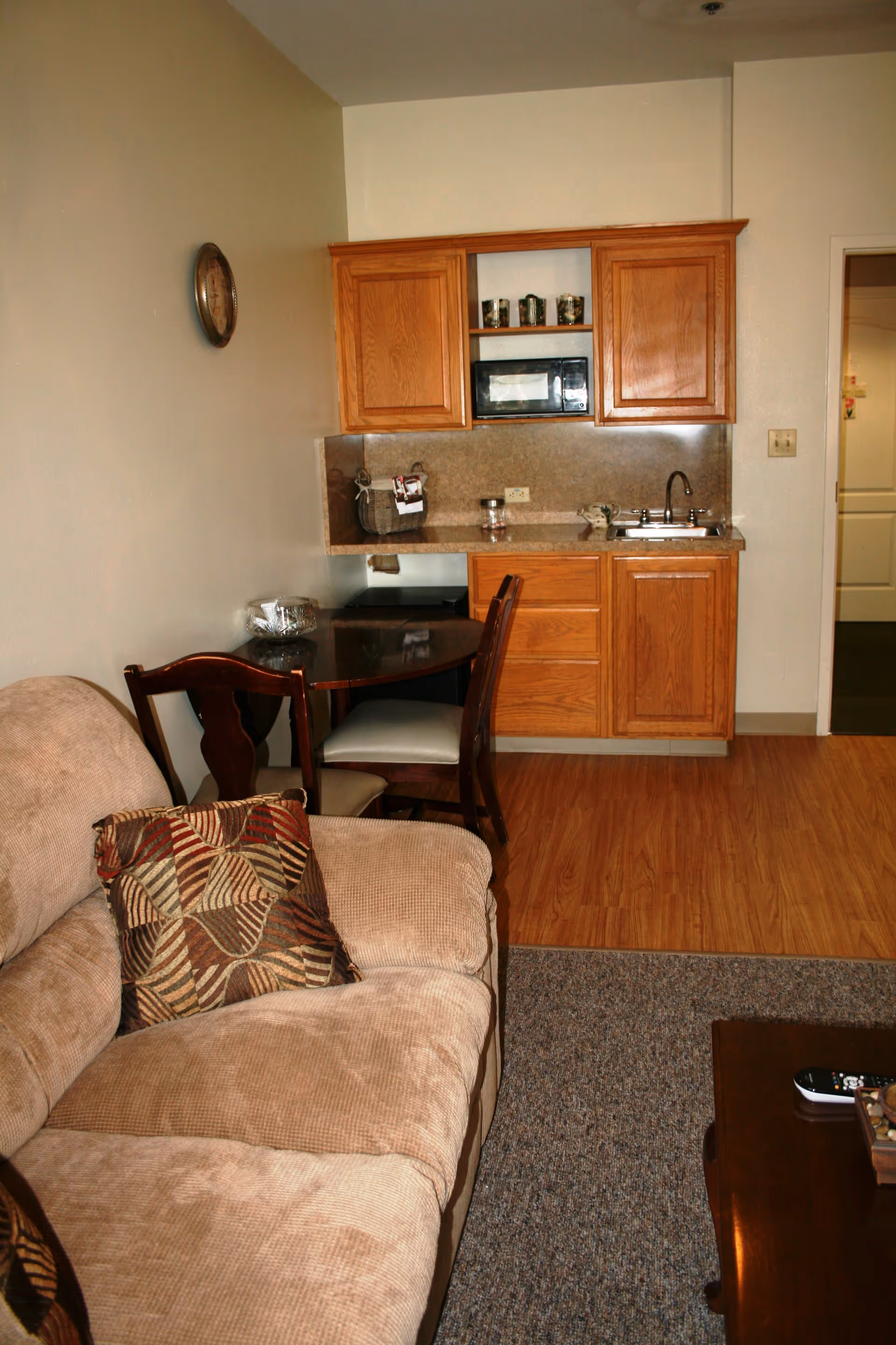 A cozy living area with a beige couch adorned with patterned cushions, a small wooden dining table with two chairs, and a kitchenette featuring wooden cabinets, a microwave, a sink, and a countertop. The floor transitions from carpet in the living area to wood in the kitchenette. A clock is mounted on the wall above the couch.