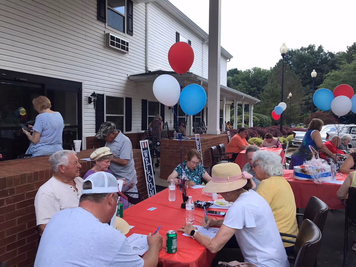 A group of elderly people seated at tables covered with red tablecloths outside a senior living facility. They are engaged in writing or filling out papers. The building has white siding with black shutters, and there are red, white, and blue balloons tied to the tables. A 'WELCOME' sign is visible near the building entrance. Trees and bushes surround the area, and there are street lamps in the background.