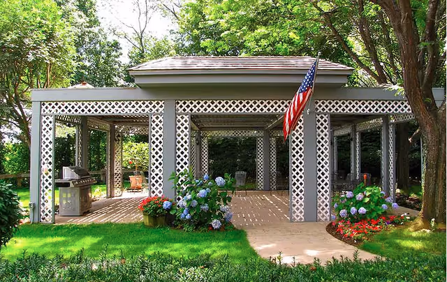 Outdoor pavilion structure with white lattice columns and a roof, surrounded by green grass, flowering plants, and trees. An American flag is hanging from one of the columns. There is a grill and outdoor seating visible under the pavilion.