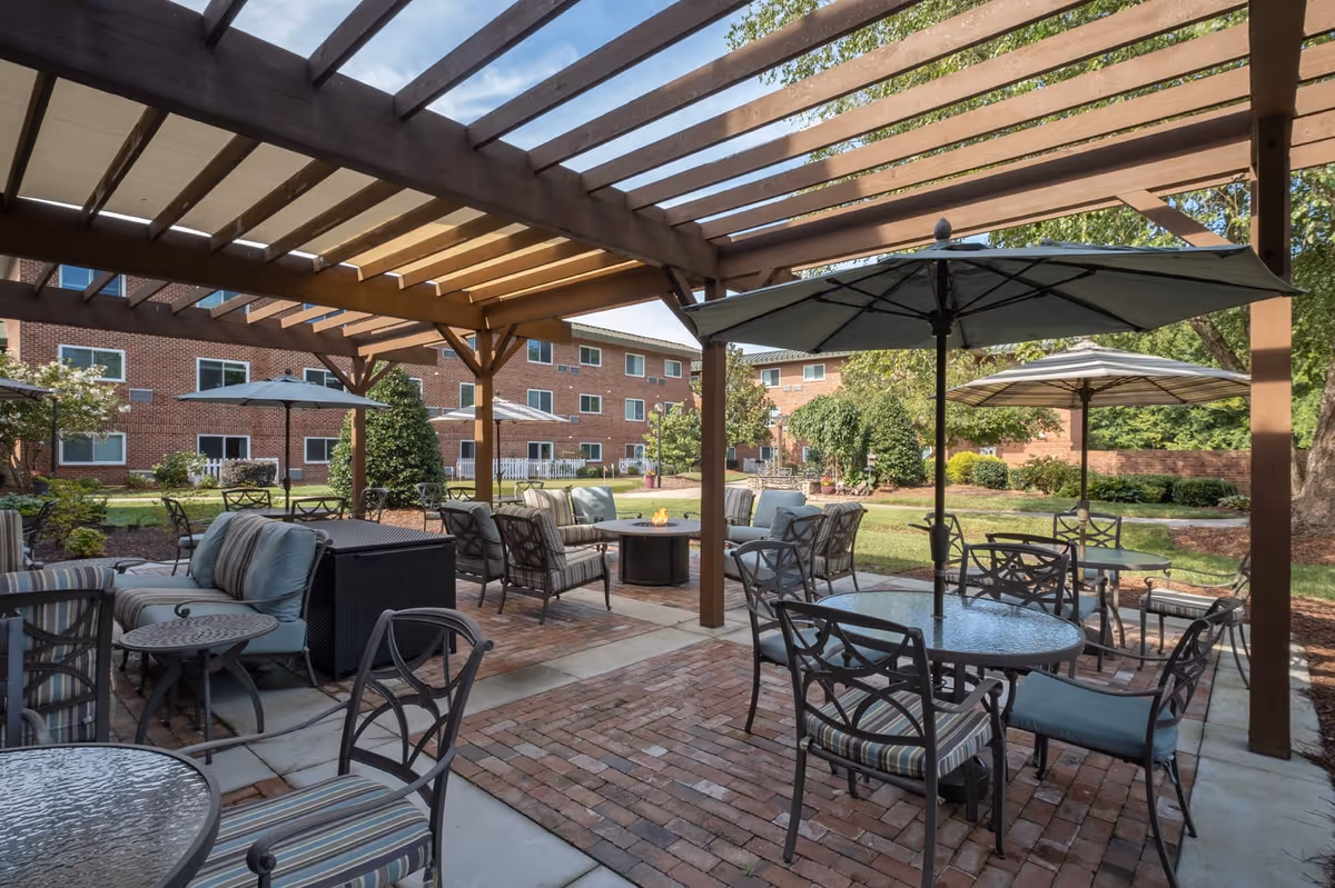 Outdoor seating area at Bartlett Reserve with multiple glass-top tables and cushioned chairs under large umbrellas and a wooden pergola. The area is paved with brick and concrete, surrounded by green grass, trees, and shrubs, with a multi-story brick building in the background.