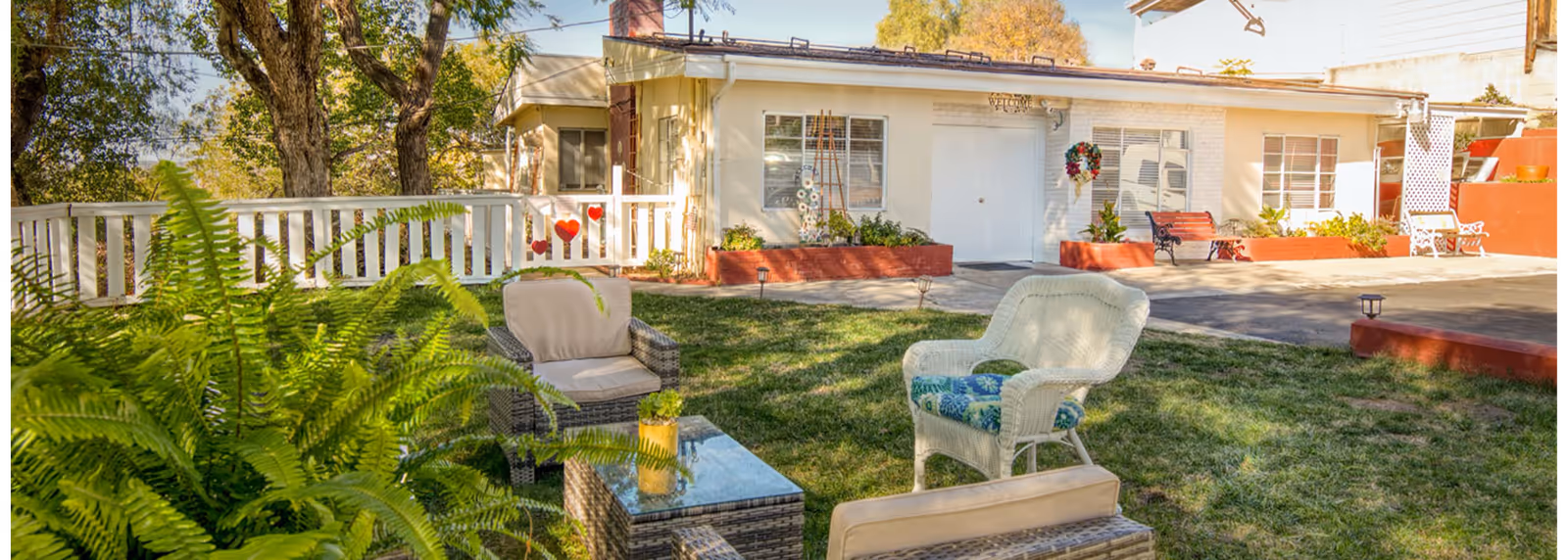 Outdoor seating area with cushioned wicker and white chairs arranged around a glass-top table on a grassy lawn in front of a single-story building with white walls and red trim. There are plants and trees surrounding the area, and a white fence with heart decorations is visible in the background.