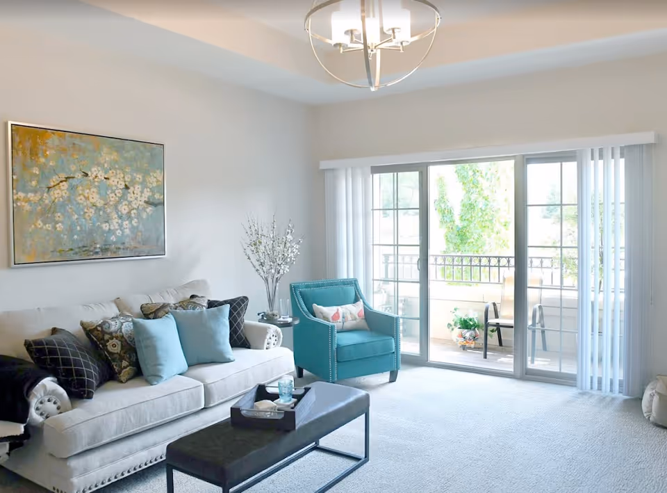 A bright living room with a beige sofa adorned with multiple decorative pillows, a teal armchair with a floral cushion, a black rectangular coffee table with a tray and glass on it, a painting of white flowers on a blue and gold background hanging on the wall, a vase with white flowers on a side table, and large sliding glass doors leading to a balcony with outdoor chairs and plants.