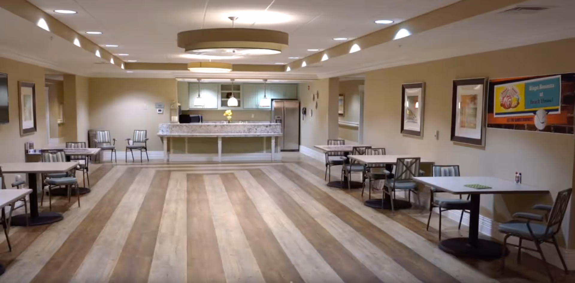 A spacious dining area with several tables and chairs arranged along the walls. The room features wood-patterned flooring and a ceiling with recessed lighting and circular light fixtures. At the far end, there is a kitchen counter with a granite countertop, pendant lights, and stainless steel appliances including a refrigerator. The walls are decorated with framed artwork and a colorful sign.