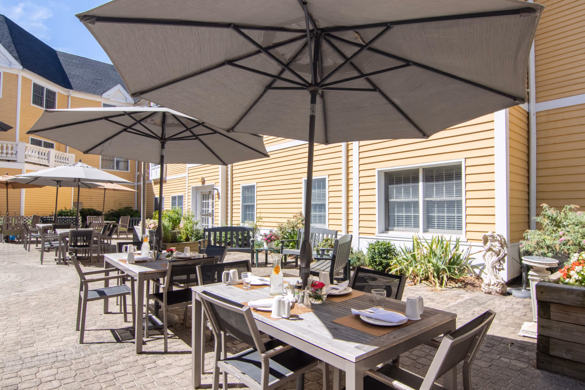Outdoor patio area at Carriage Green at Milford with multiple tables and chairs under large umbrellas. Tables are set with placemats, plates, cups, and a pitcher of water. The patio is paved with stone tiles and surrounded by yellow siding building walls, windows, and some greenery including plants and flowers.