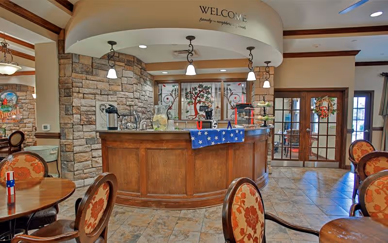 Interior view of a welcoming reception area in a senior living facility with a curved wooden desk, stone accent wall, hanging pendant lights, and decorative elements including a blue star-spangled cloth and a glass container with lemonade. There are round tables and chairs with floral upholstery in the foreground, and double glass doors with a wreath in the background.