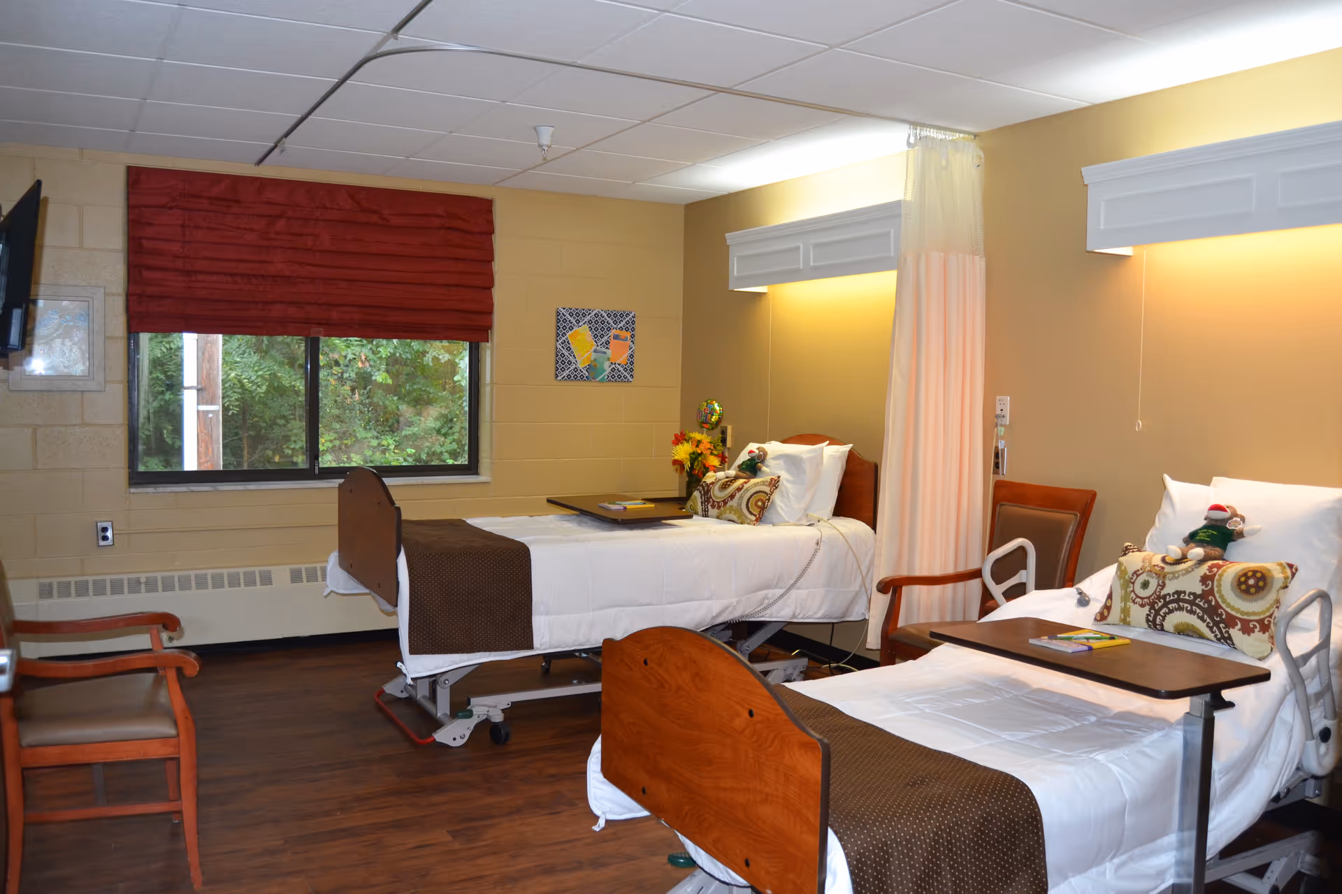 A room with two hospital-style beds, each with white bedding, brown blankets, and decorative pillows. There are overbed tables on each bed, a wooden chair between the beds, a window with a red curtain, and a wall-mounted TV. The walls are beige, and the floor is wooden. A privacy curtain is partially drawn between the beds.