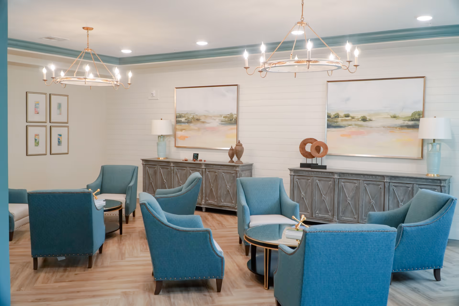 A well-lit lounge area with six teal upholstered armchairs arranged around two circular glass-top tables. The room features light wood flooring, two large landscape paintings on the white shiplap wall, two gray wooden sideboards with decorative items and lamps, and two large chandeliers hanging from the ceiling.