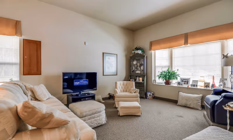 A cozy living room with a beige sofa, a cushioned armchair with an ottoman, a flat-screen TV on a stand, a tall wooden cabinet, and a large window with blinds and an orange valance. The window sill holds several framed photos, a potted plant, and decorative items. The room has neutral-colored walls and carpeted flooring.