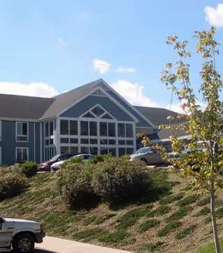 Exterior view of a multi-story senior living facility building with large windows and a sloped roof, surrounded by a landscaped area with bushes, a tree, and parked cars under a blue sky with some clouds.