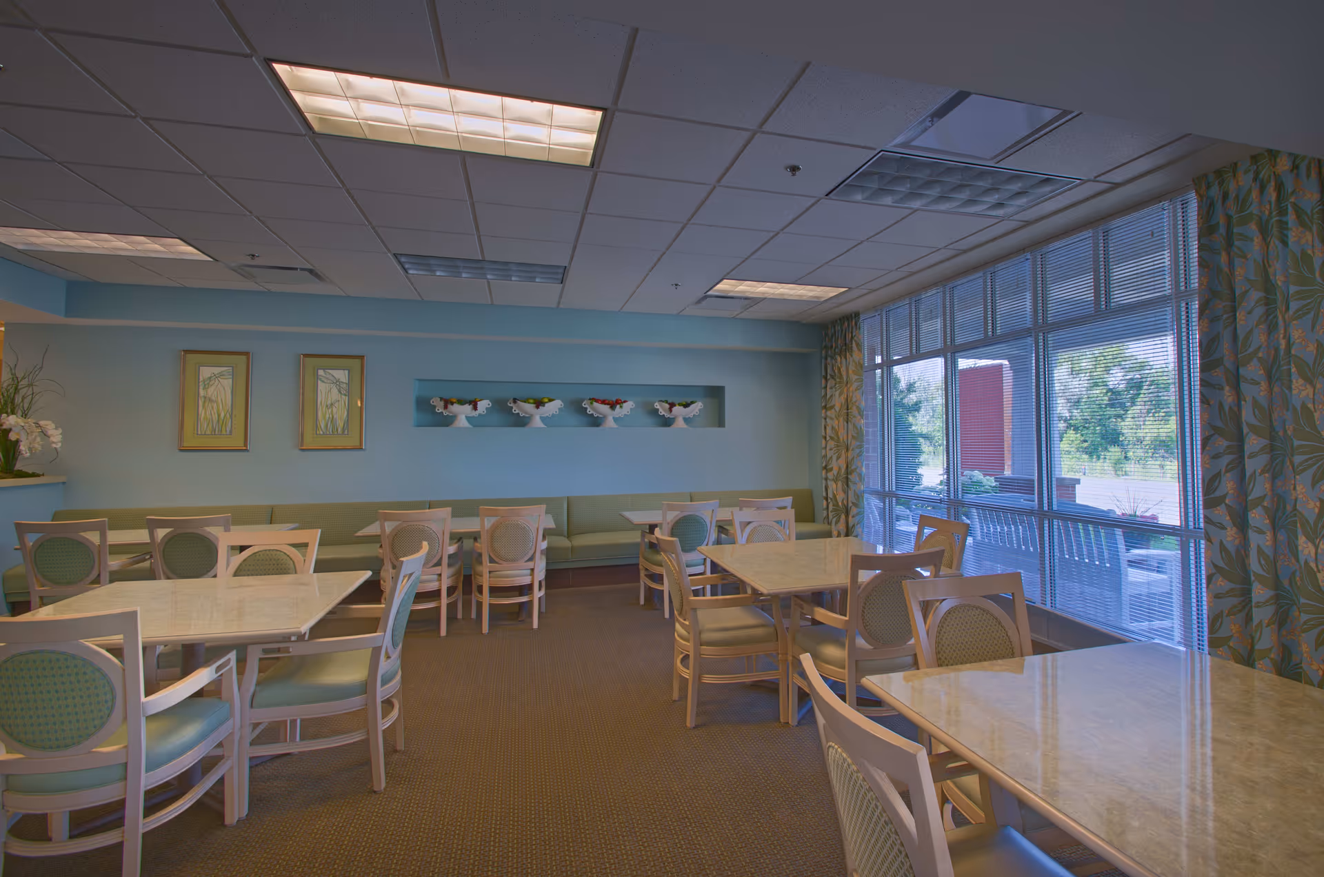 A dining room with several tables and chairs arranged neatly. The walls are painted light blue with framed artwork and decorative vases on a recessed shelf. Large windows with blinds and floral curtains allow natural light to enter the room.