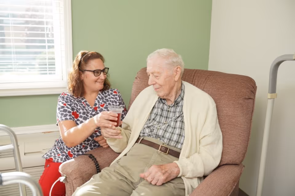 A female caregiver with glasses and a patterned shirt is smiling and handing a drink to an elderly man sitting in a comfortable chair inside a room with light green walls and a window with blinds.