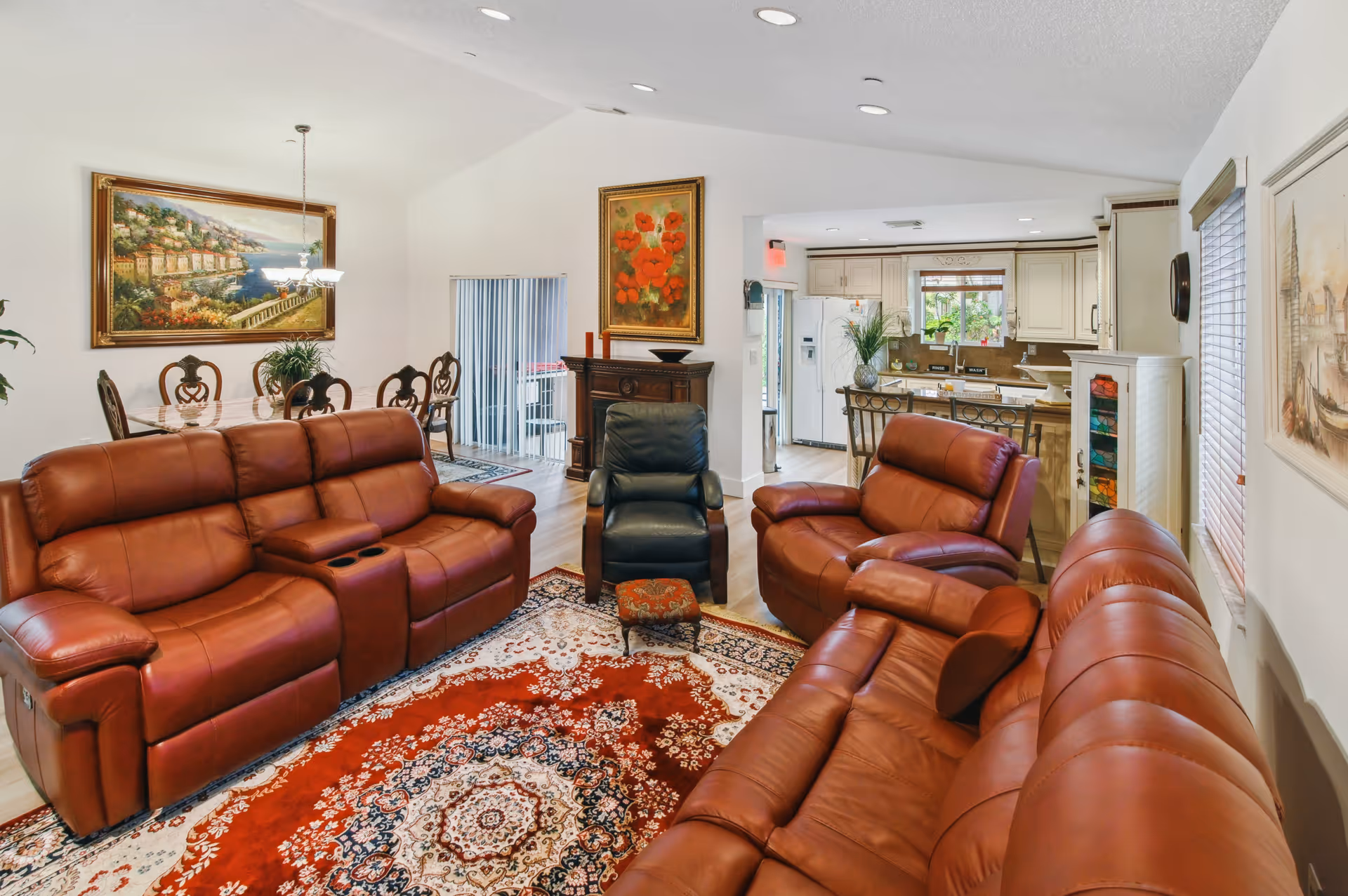 A spacious living room with three brown leather sofas and one black armchair arranged around a large ornate red and white area rug. Behind the seating area, there is a dining table with six chairs and a large framed painting on the wall. To the right, an open kitchen with white cabinetry and a window overlooking greenery is visible. The room is well-lit with natural light coming through windows with blinds.