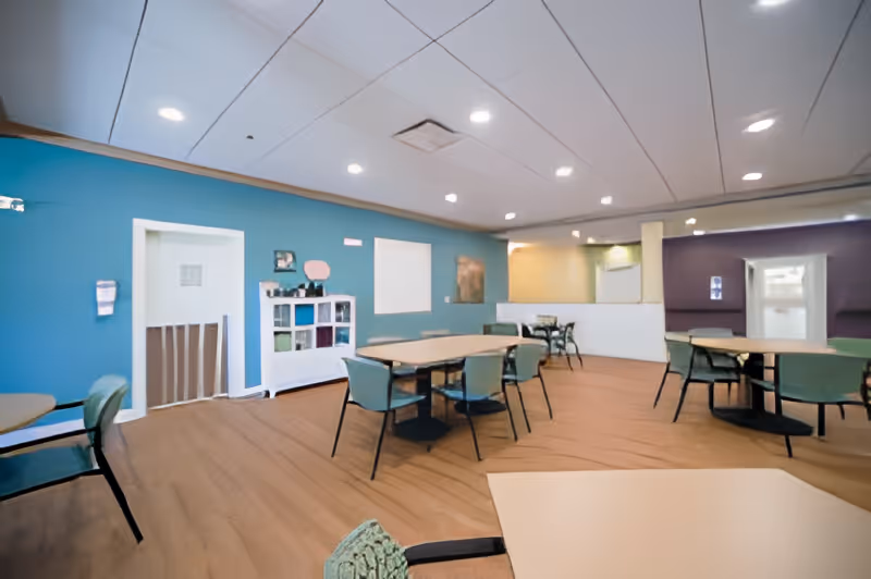 A spacious dining area with multiple tables and chairs arranged on a wooden floor. The walls are painted in blue, yellow, and purple tones. There is a white shelving unit against the blue wall with various items on it. The ceiling has recessed lighting.