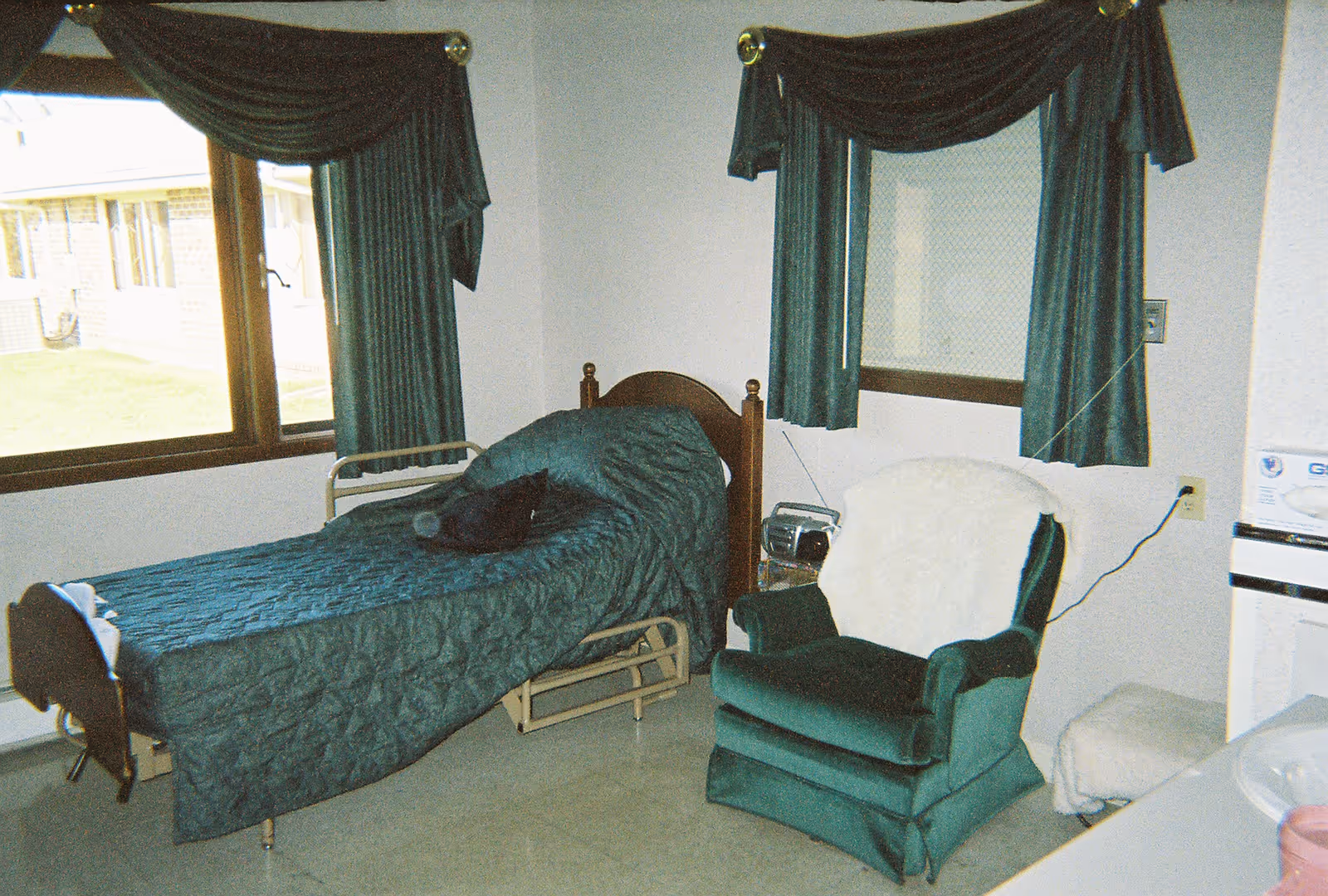 A small bedroom with a single bed covered in a dark green quilted bedspread. Next to the bed is a green upholstered armchair with a white throw draped over the back. The room has two windows with dark green curtains, and there is a small radio on a table beside the chair. The floor is light-colored, and part of a sink and countertop is visible on the right side of the image.