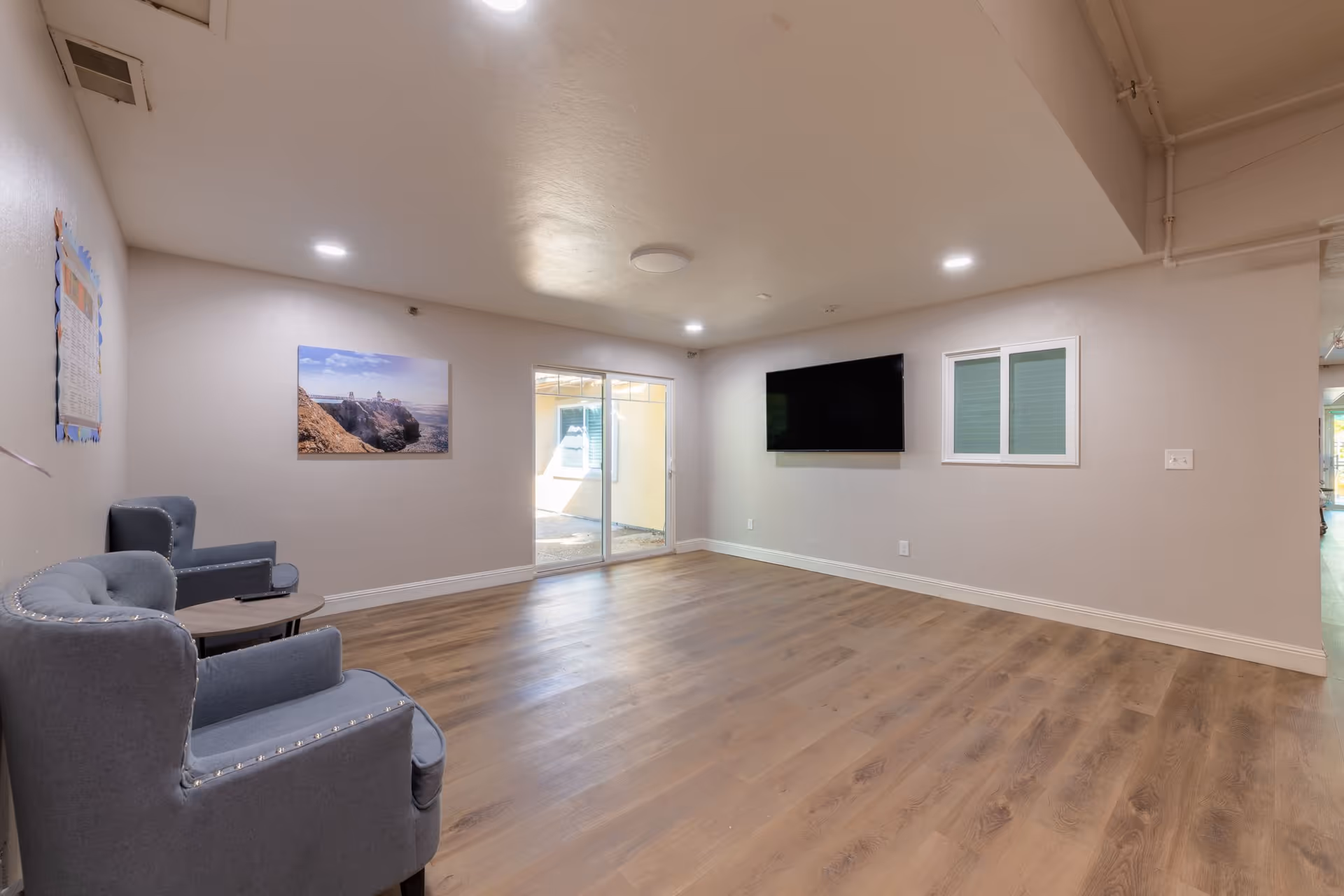 A spacious room with light wood flooring, two gray upholstered armchairs with nailhead trim on the left, a small round table between them, a large flat-screen TV mounted on the wall to the right, a sliding glass door leading to an outdoor area, and a landscape photograph hanging on the left wall.