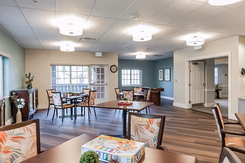 A bright and spacious common area in a senior living facility with several tables and chairs arranged for socializing or activities. The room features wood flooring, light-colored walls, ceiling lights, and large windows allowing natural light. A board game is visible on one of the tables, and there is a pool table in the background. Doors and exit signs are also visible.