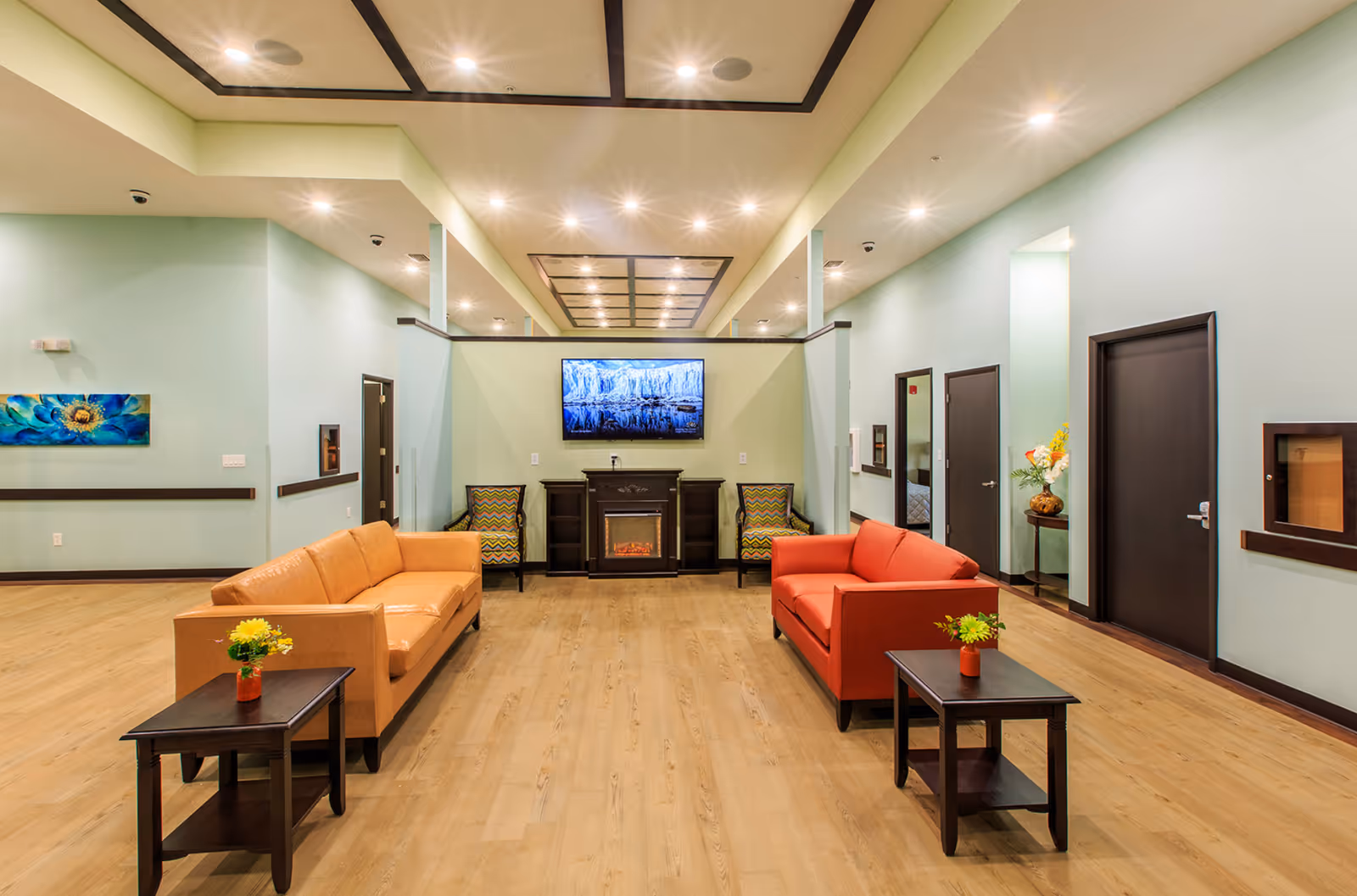 A spacious and well-lit living room area with light green walls and wooden flooring. The room features two sofas, one orange and one red, facing each other with dark wooden side tables holding small flower vases. At the far end, there is a wall-mounted flat-screen TV above an electric fireplace flanked by two patterned armchairs. Several doors and a small table with a flower arrangement are visible along the walls.