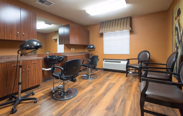Interior of a salon room with two black salon chairs in front of a counter with a sink and cabinets. There are two hair dryers on stands, a window with blinds and a valance, and three chairs lined up against the wall. The room has wood flooring and warm-toned walls.