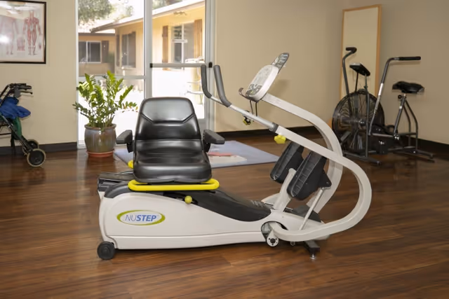 Recumbent NuStep exercise machine in a rehabilitation facility exercise room with hardwood floors, windows, a potted plant, and additional cardio equipment.